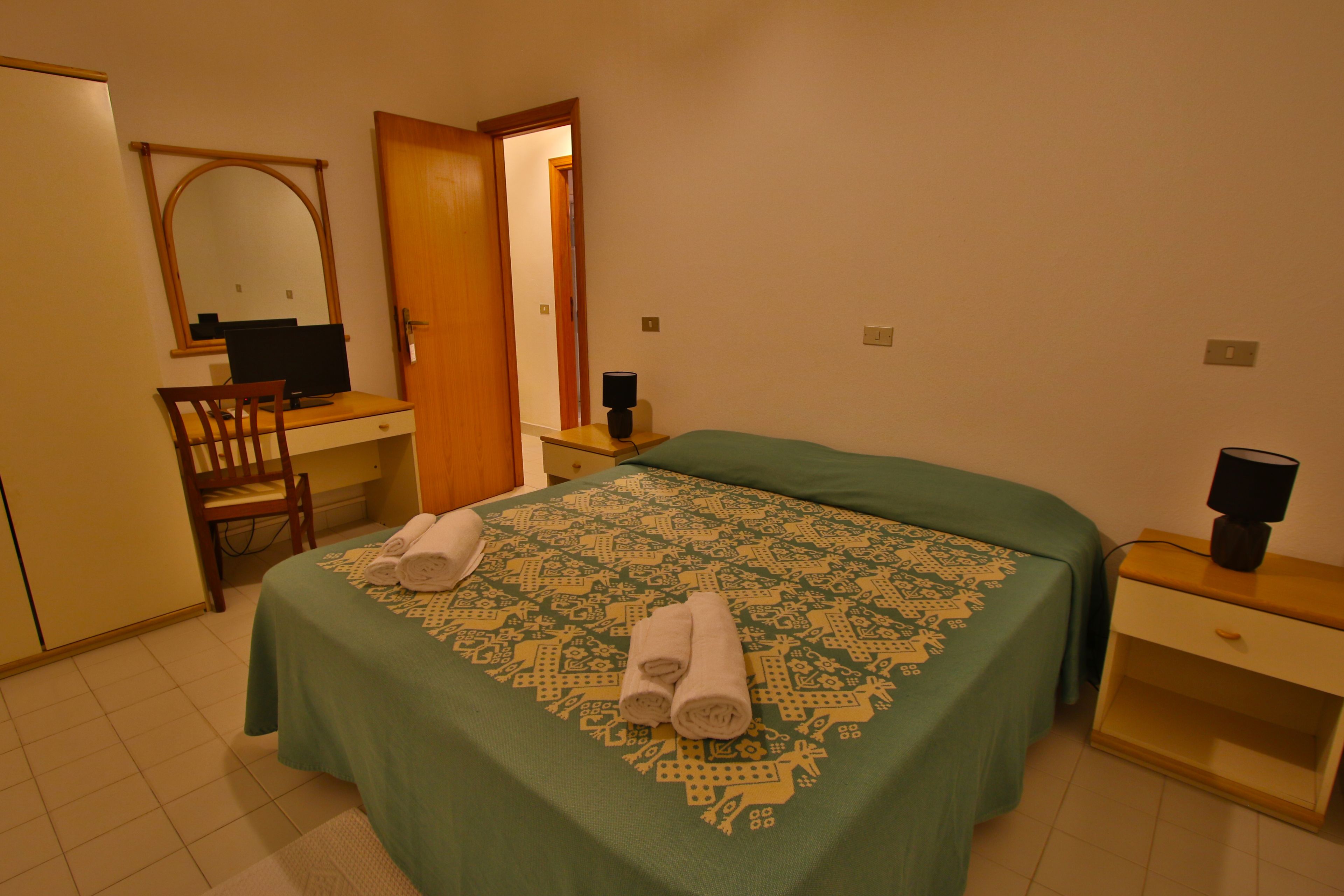 A hotel room with a bed, a desk, a chair, a nightstand, and a mirror.