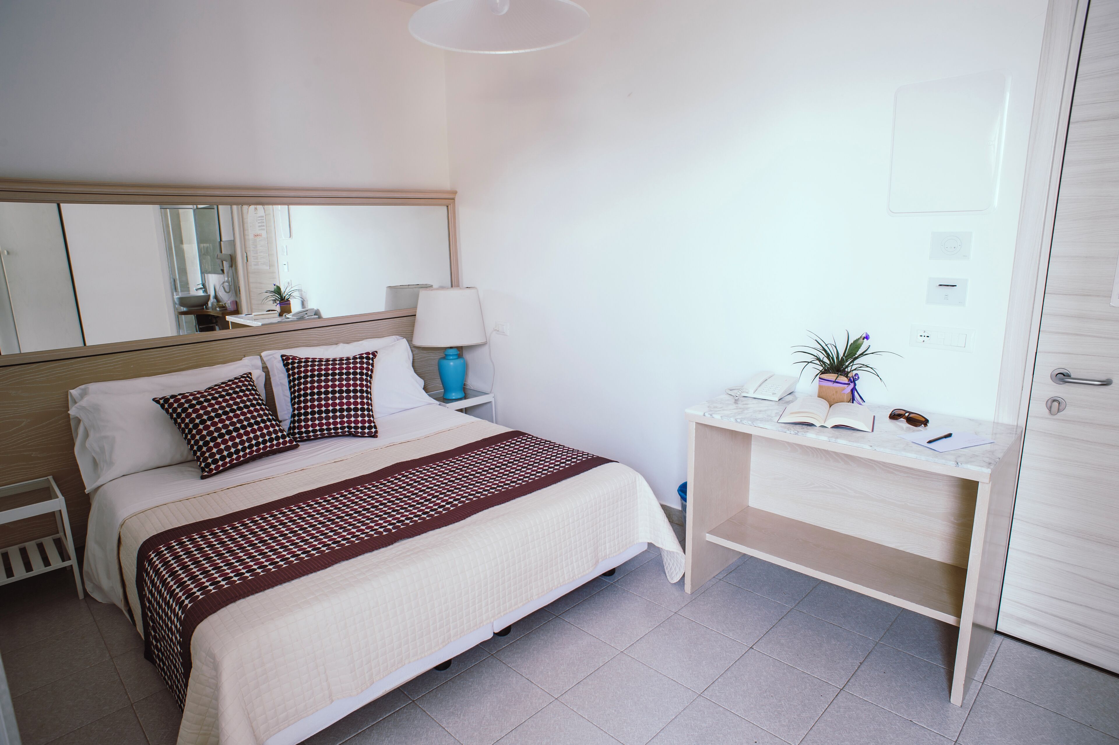 A neatly arranged bedroom with a bed, bedside table, lamp, and a desk with a chair.