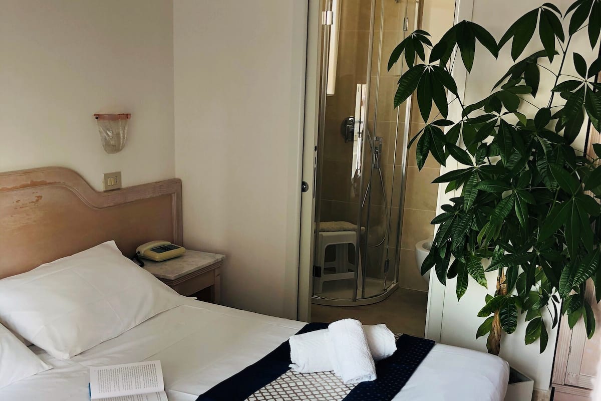 Une chambre d’hôtel avec un lit bien fait, une table de chevet avec une lampe, un espace douche et une plante en pot.