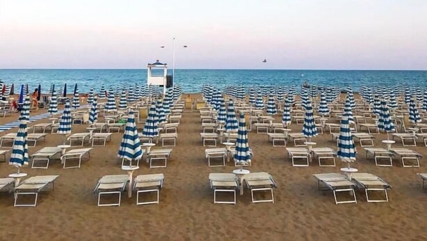 A beach with rows of sun loungers and striped umbrellas, a lifeguard tower, and the ocean in the background.