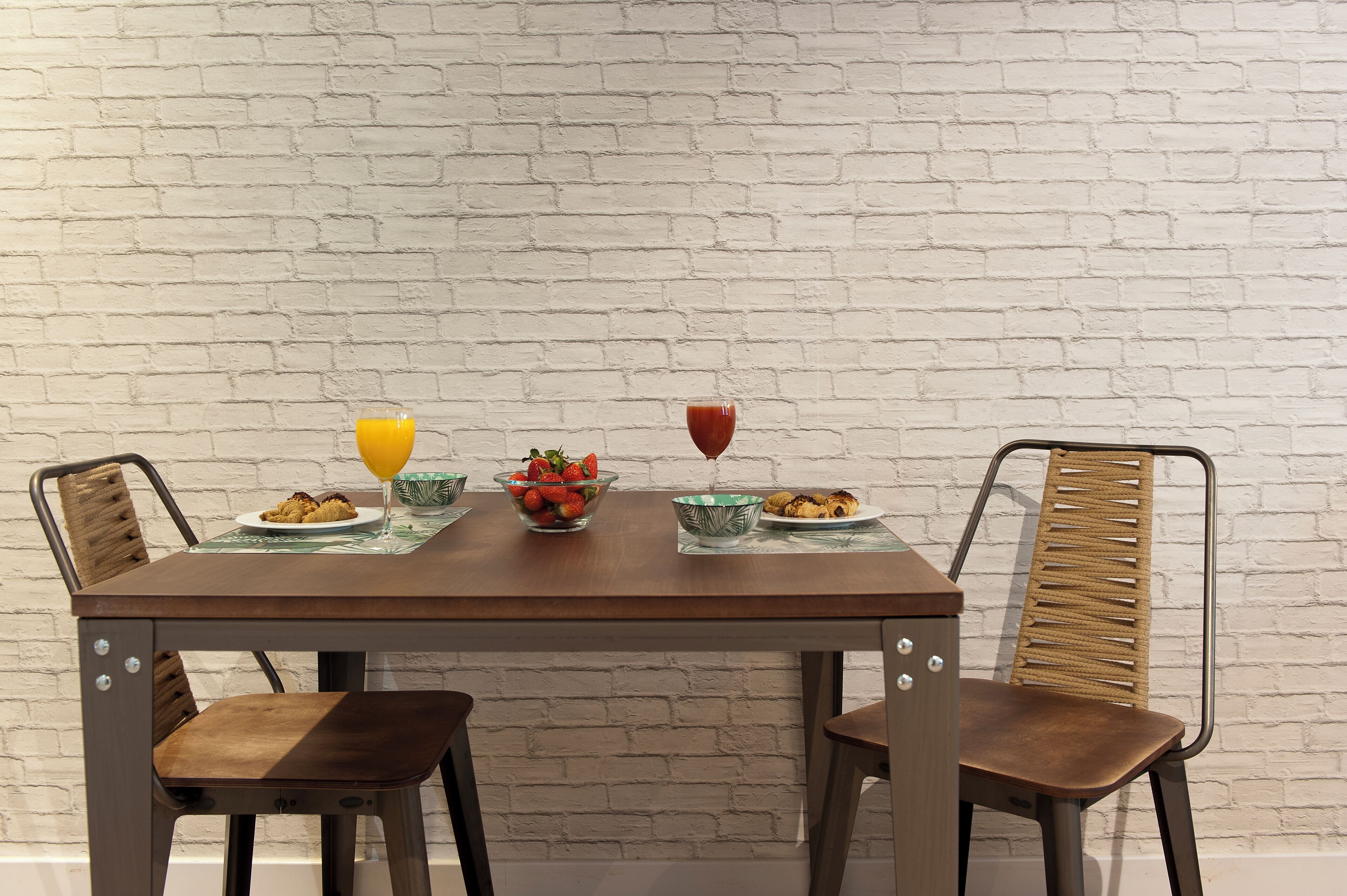 A dining table with two chairs, a white brick wall, and a bowl of fruit.