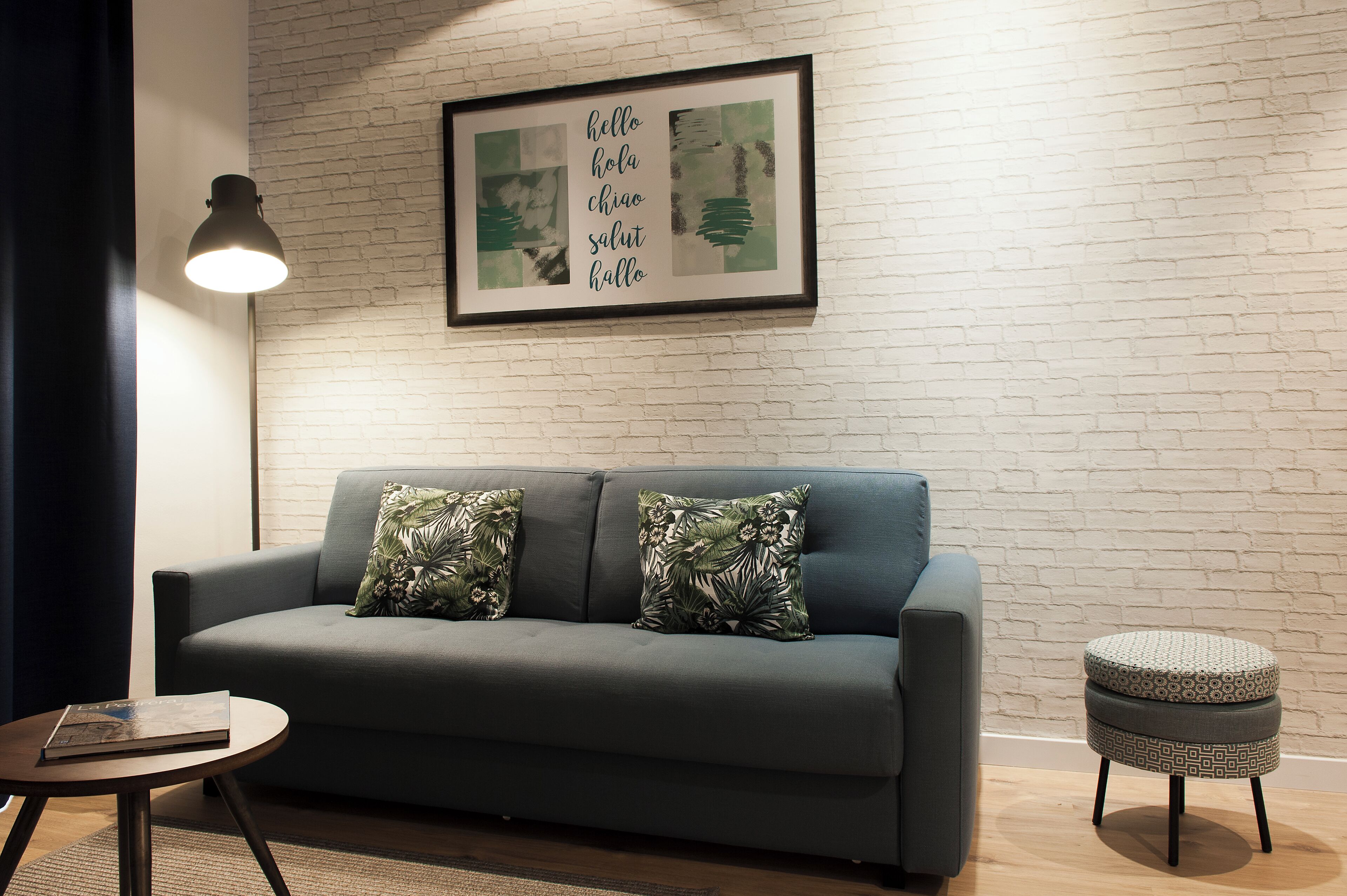 Un salón moderno con un sofá gris, dos cojines estampados, un taburete redondo pequeño, una obra de arte enmarcada con saludo en varios idiomas y una lámpara de pie.