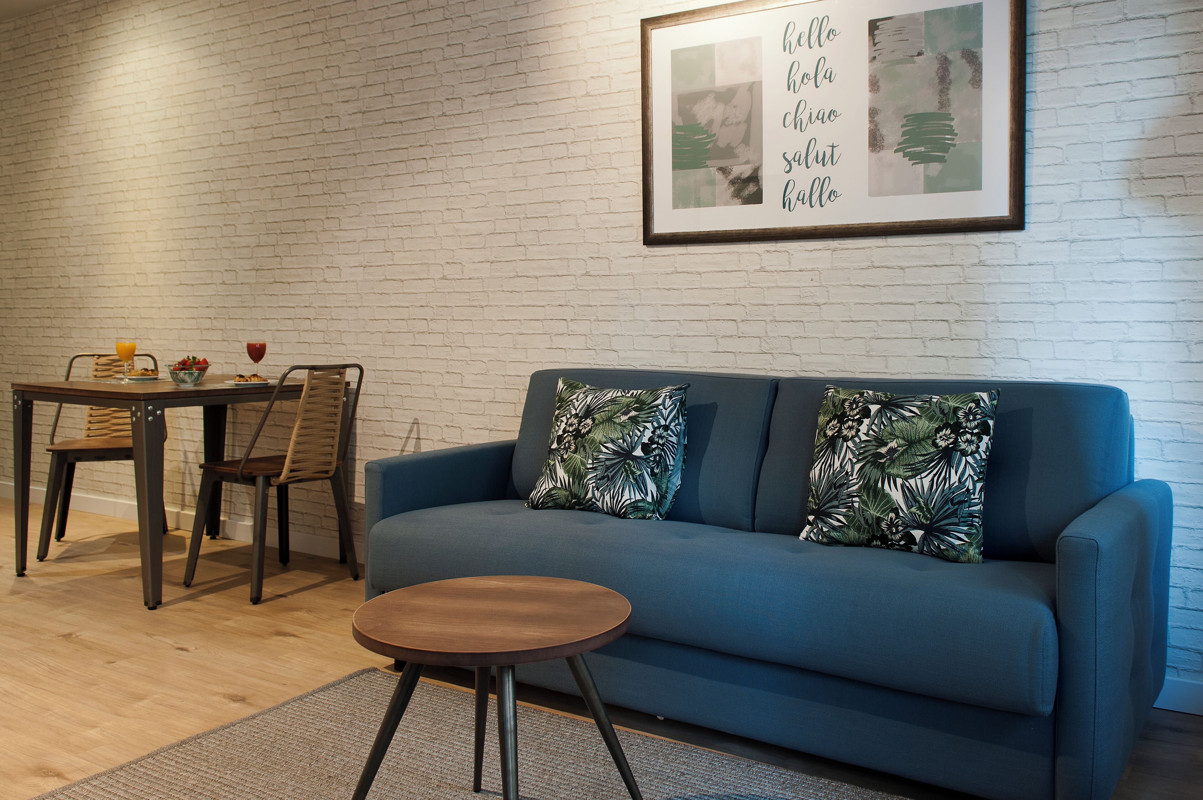Una sala de estar con un sofá azul, una mesa de centro de madera, una mesa de comedor con sillas y una pared de ladrillo blanco.