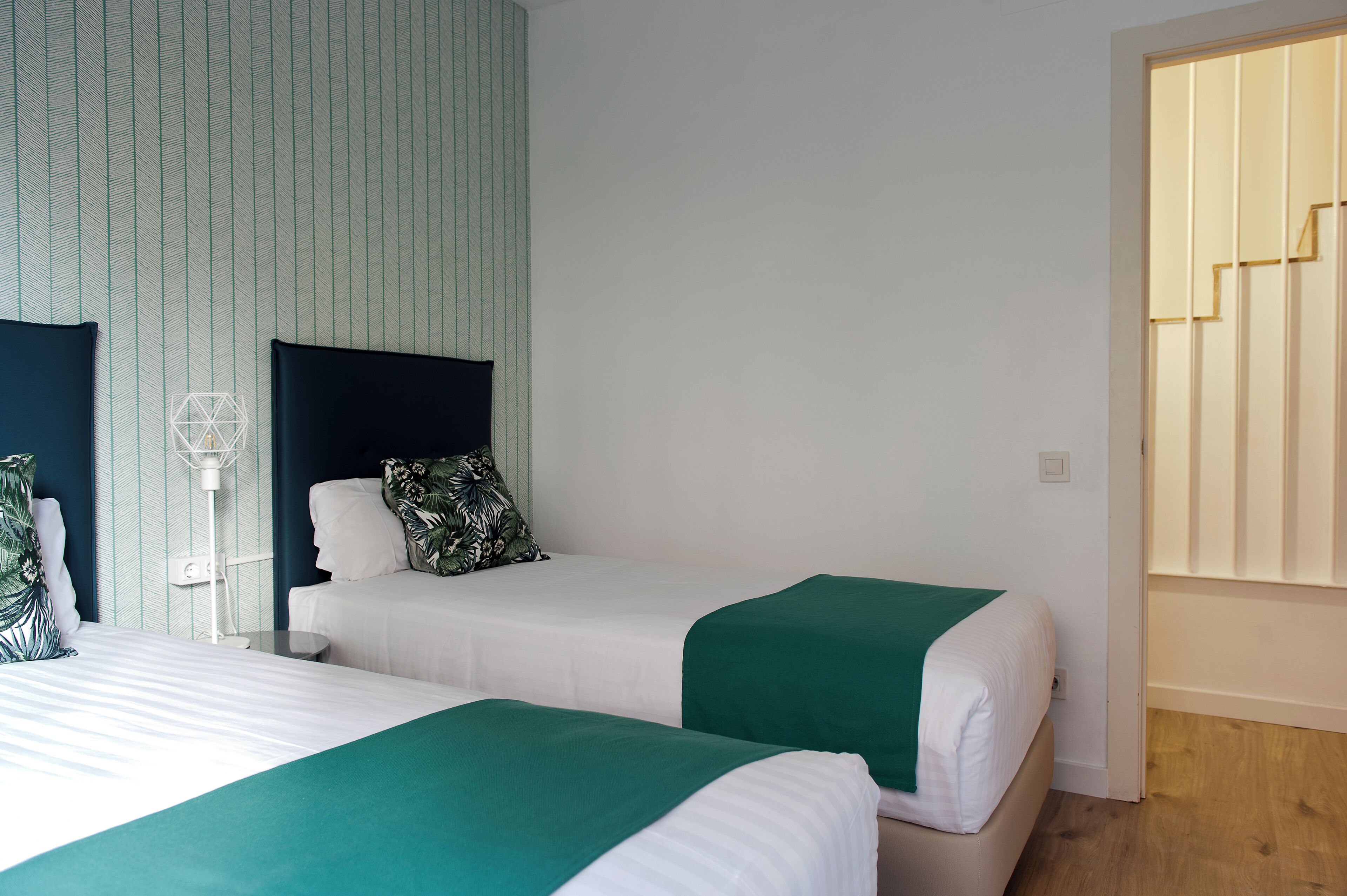 A hotel room with two beds, each with a green bedspread, white linens, and a dark headboard. A white wall-mounted lamp is visible, and the room has a wooden floor.