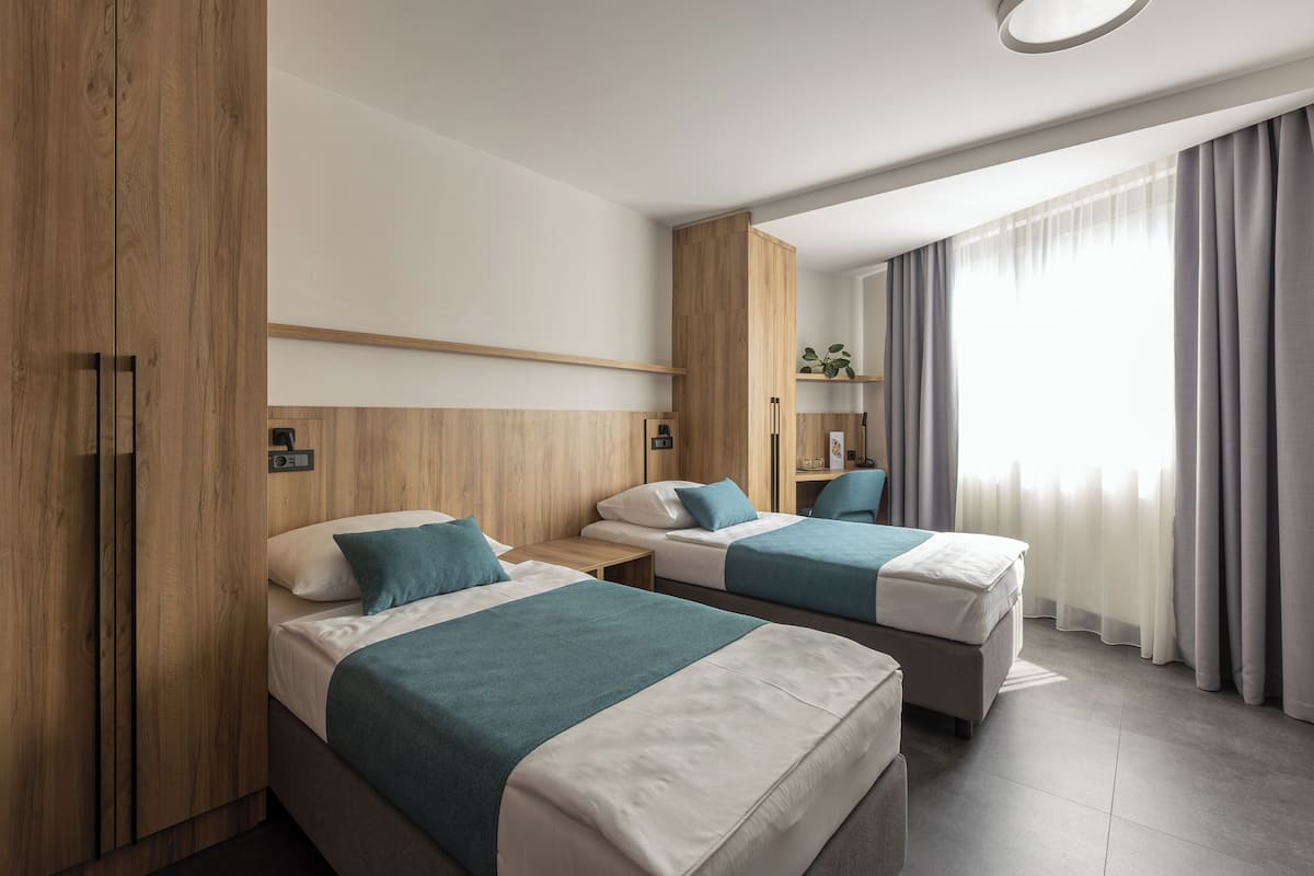 A hotel room with two beds, wooden wardrobe, a small table, and a window with curtains.