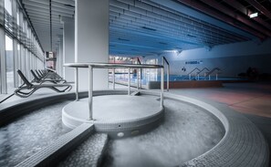 A modern indoor swimming pool with a central circular platform, surrounded by seating areas and a tiled floor. - Hotel Zdravilisce Lasko (Lasko)