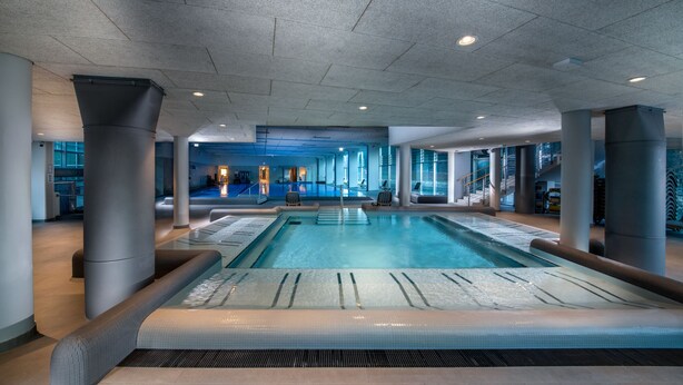 Ein großer, überdachter Schwimmbereich mit klarem Wasser, umgeben von Säulen und modernem Architekturdesign.