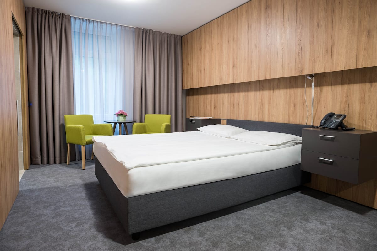 A hotel room with a large bed, two chairs, a small table with a vase, and a wooden wardrobe.