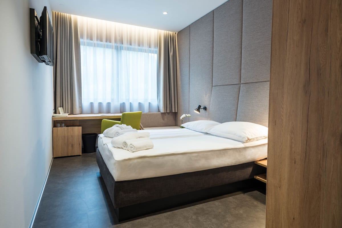 A modern hotel room with a large bed, a desk with a chair, a TV mounted on the wall, and a window with curtains.