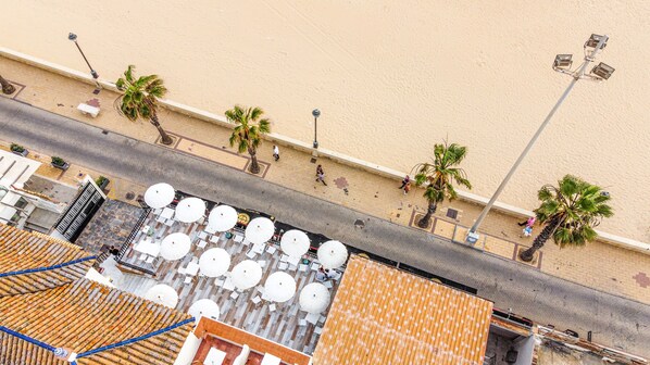 An aerial view of a beachside area with a row of white umbrellas and chairs, a sandy beach, and palm trees. - Hotel Playa De Regla (Chipiona)