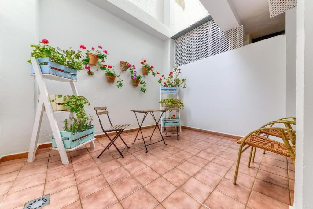 Une petite terrasse avec un mur blanc, un sol en terre cuite et diverses plantes en pot. On y trouve deux chaises et une petite table.