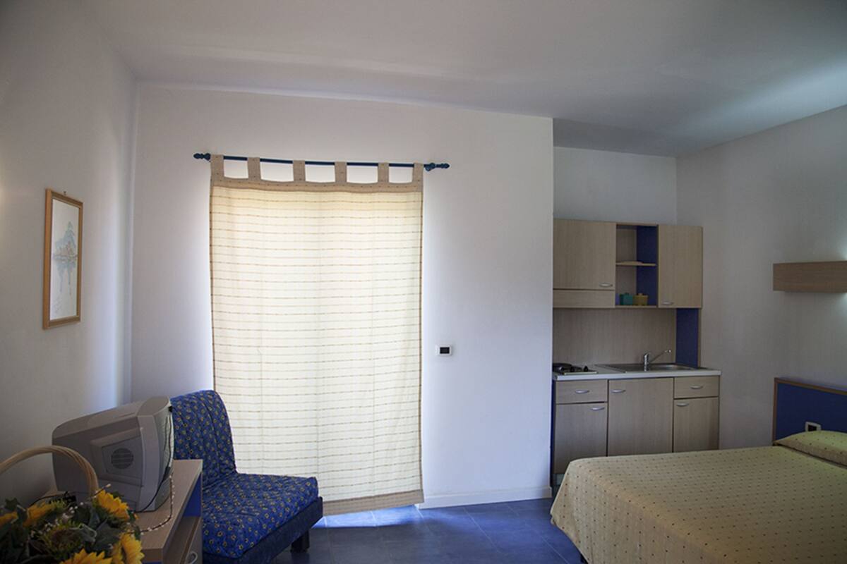 Een slaapkamer met een bed, een bureau met computer, een stoel, een raam met jaloezieën, een kitchenette en een badkamer.