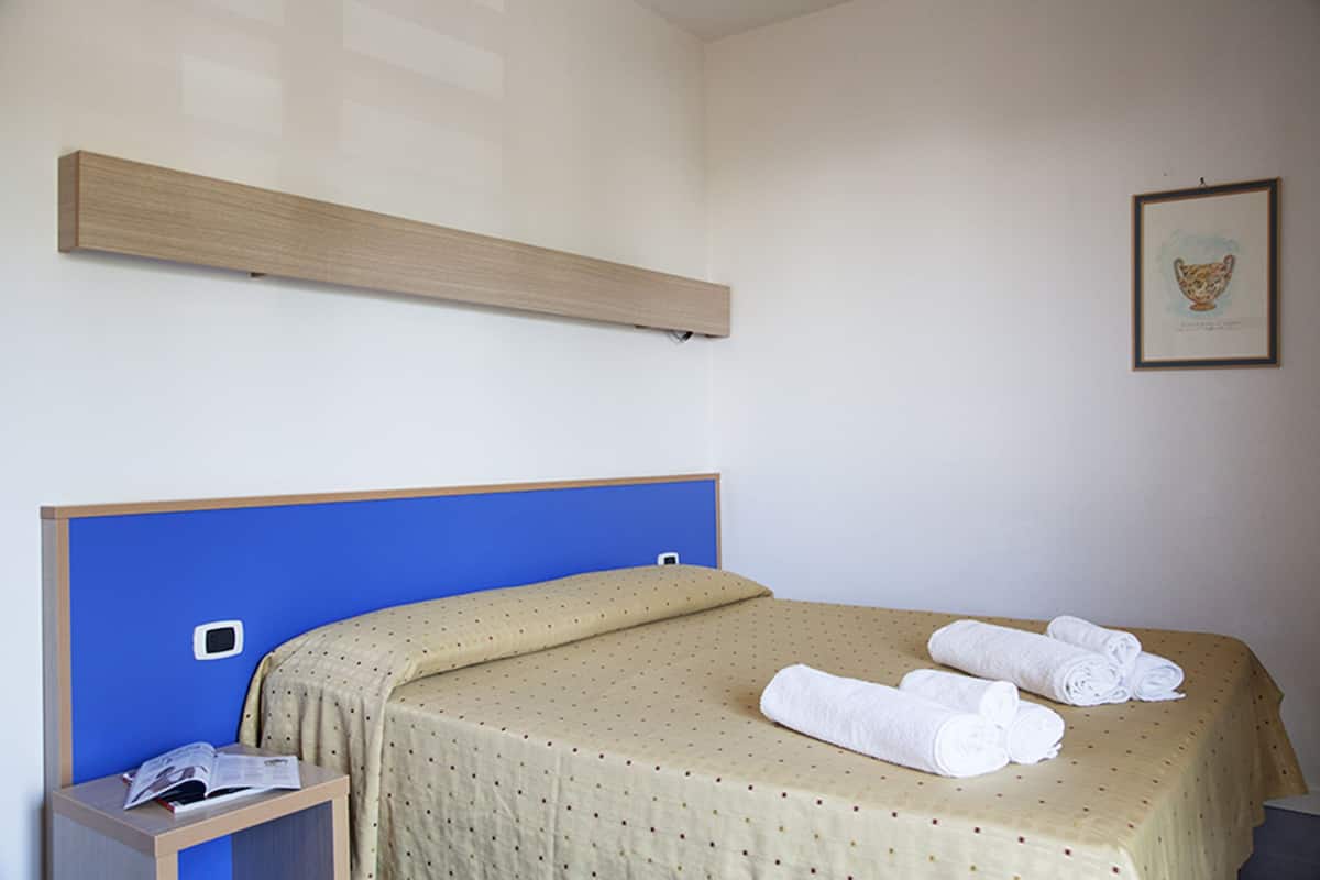Een hotelkamer met een bed, een nachtkastje, een ingelijste foto aan de muur en een houten plank boven het bed.