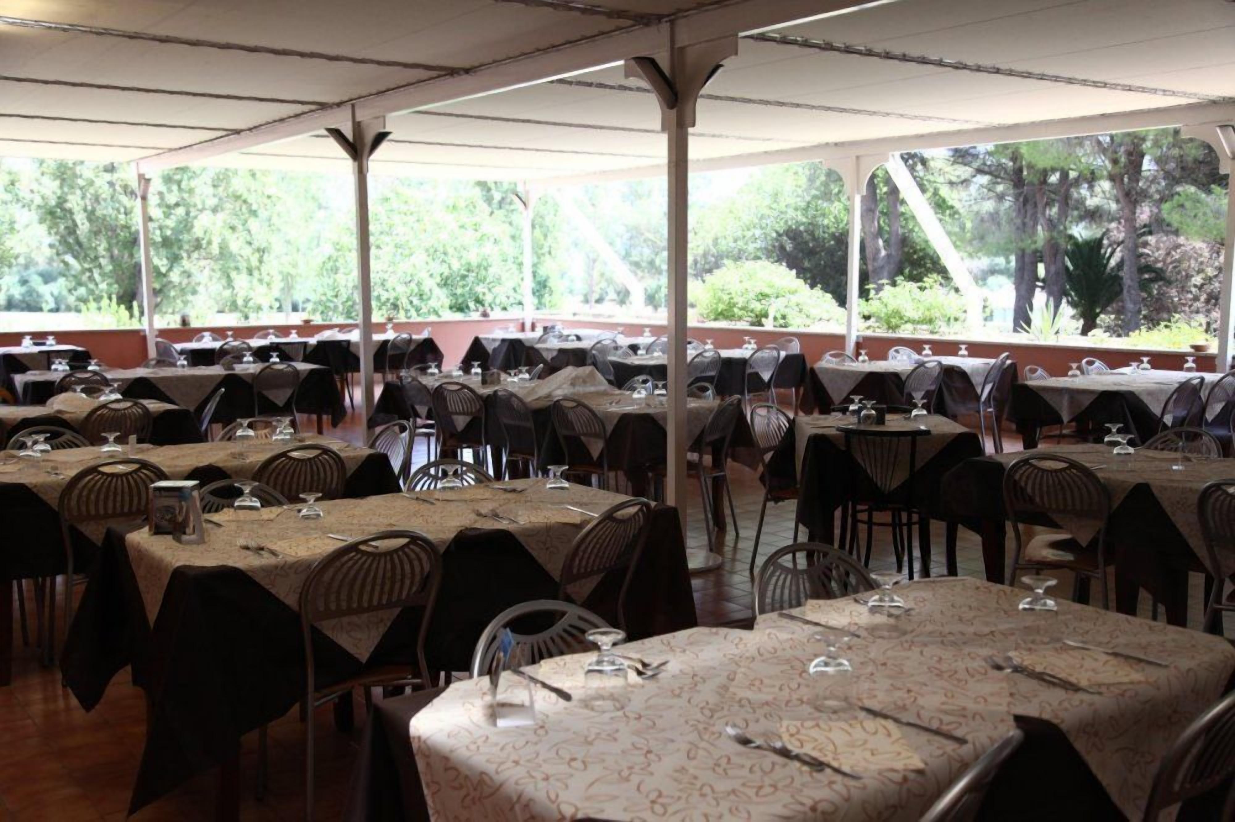 A covered outdoor dining area with multiple tables and chairs, set against a backdrop of trees.