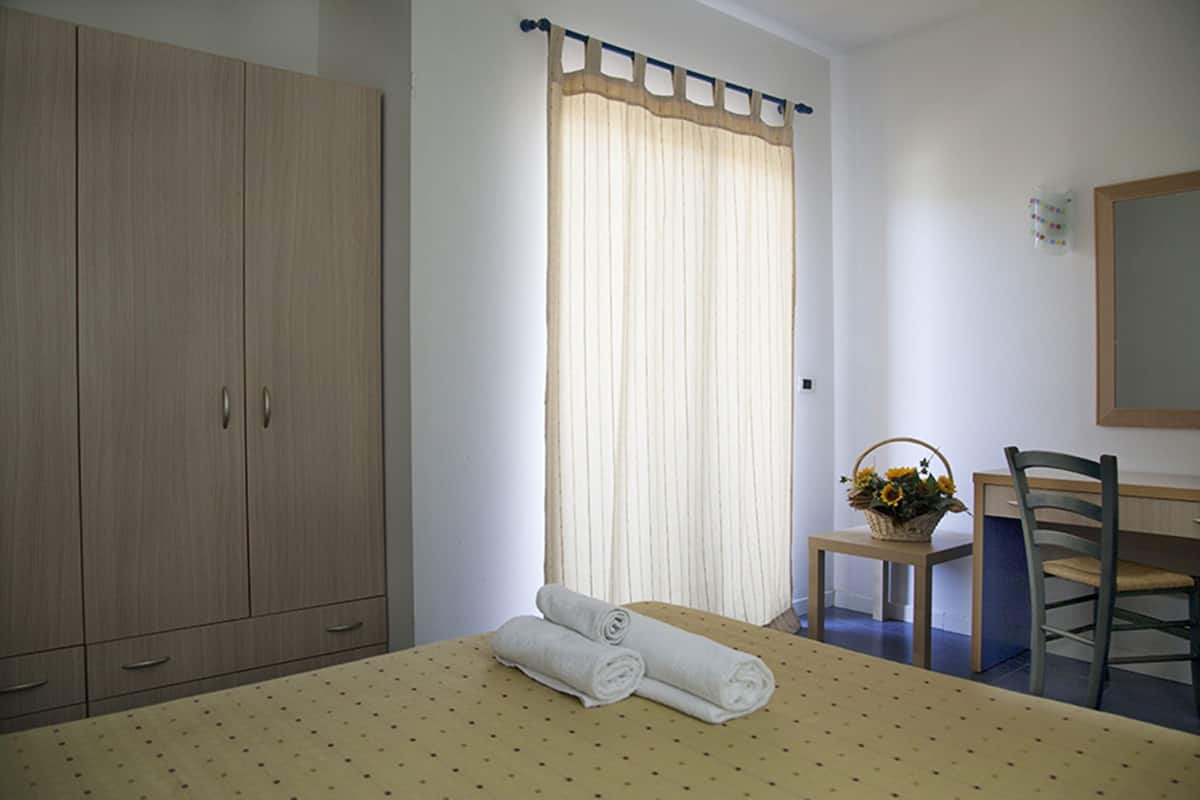 Een slaapkamer met een bed, een houten garderobe, een raam met gordijnen, een kleine tafel met een bloemenmandje en een stoel.