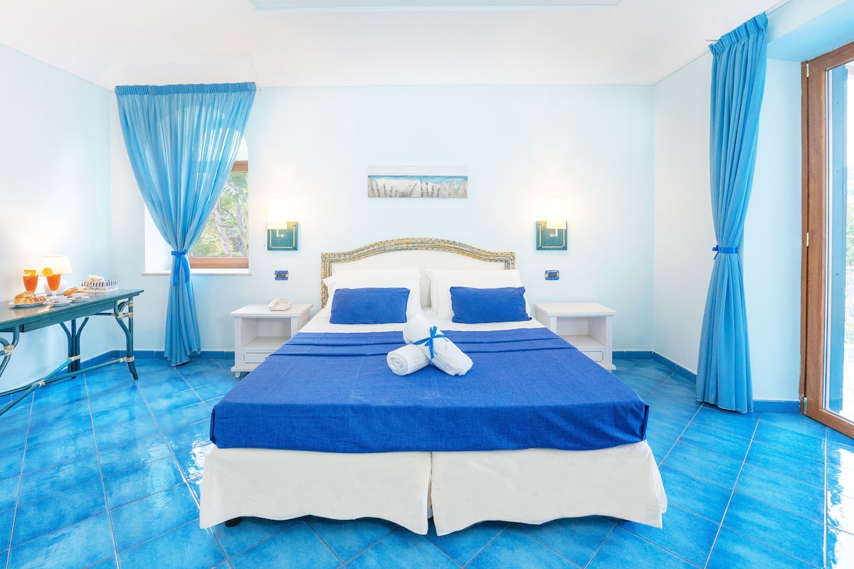 A hotel room with a large bed, two bedside tables, a small table with a lamp, and a window with curtains.