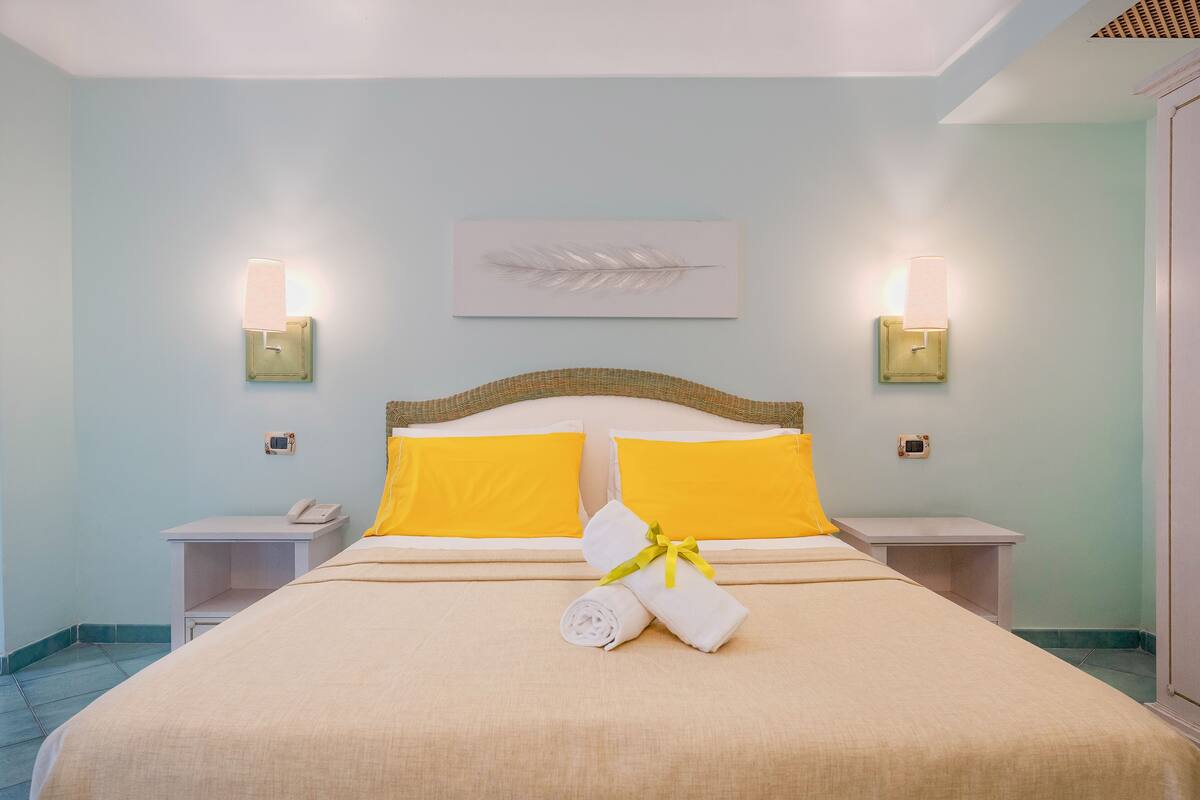 A neatly made bed with yellow pillows, a white towel, and a feather artwork on the wall.