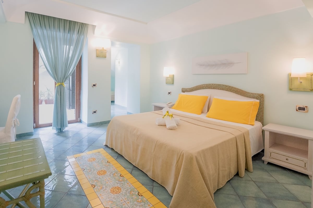 A hotel room with a bed, bedside tables, a chair, a small table, and a window with curtains.