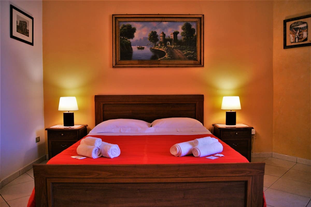 A hotel room with a wooden bed, red bedspread, two nightstands with lamps, and a painting on the wall.