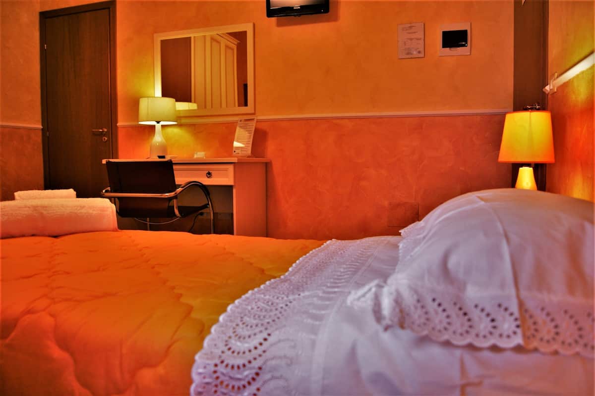 A hotel room with a bed, a desk with a chair, a lamp, a mirror, and a door.