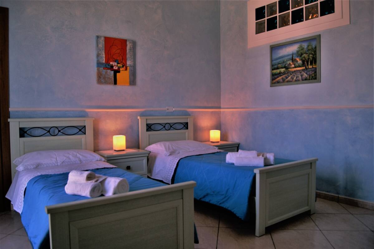 A room with two beds, each with a blue bedspread and white pillows, and two bedside lamps.