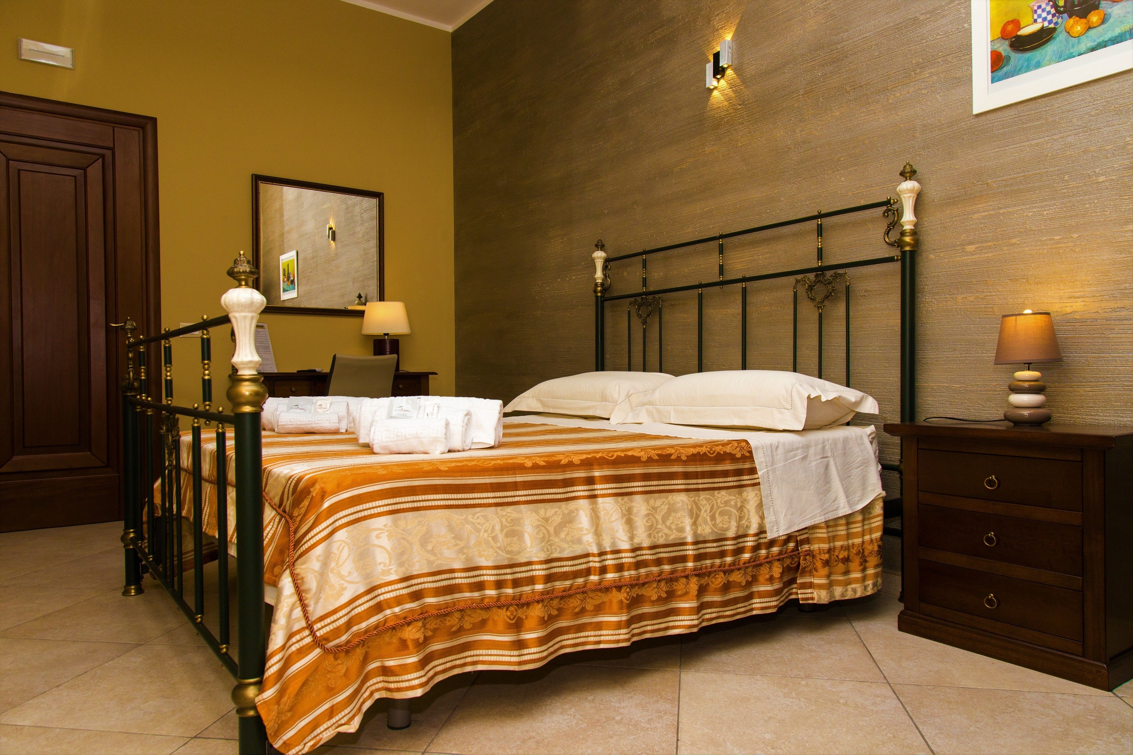 Cama bem arrumada com colcha listrada, mesa de cabeceira de madeira com abajur e um quadro na parede.
