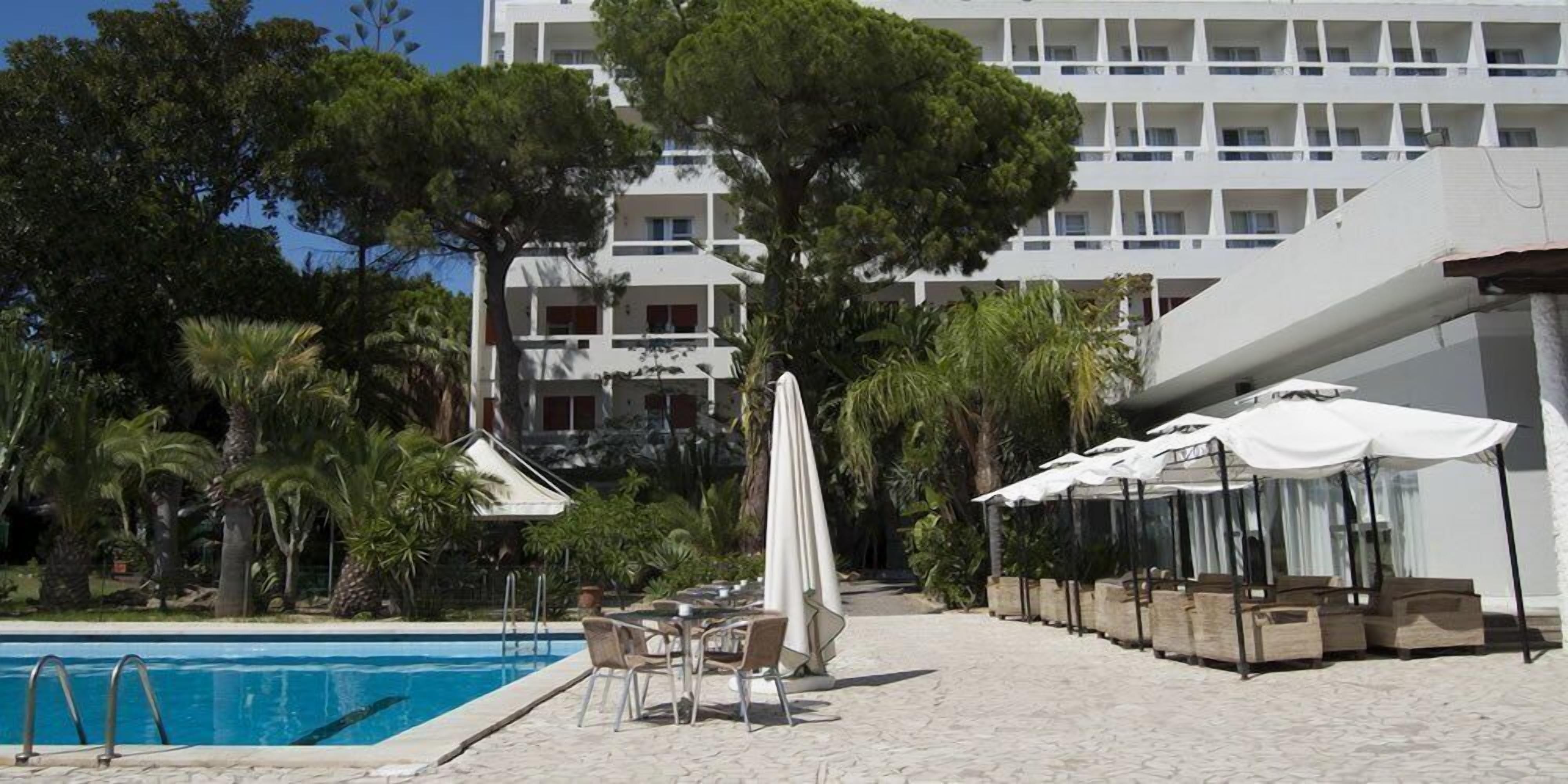 Un espace piscine doté de chaises longues et de parasols, entouré de palmiers et avec un immeuble à plusieurs étages en arrière-plan.