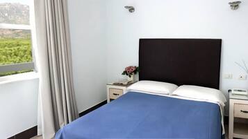 A hotel room with a bed, a nightstand, a bedside table, and a window with a view of greenery.