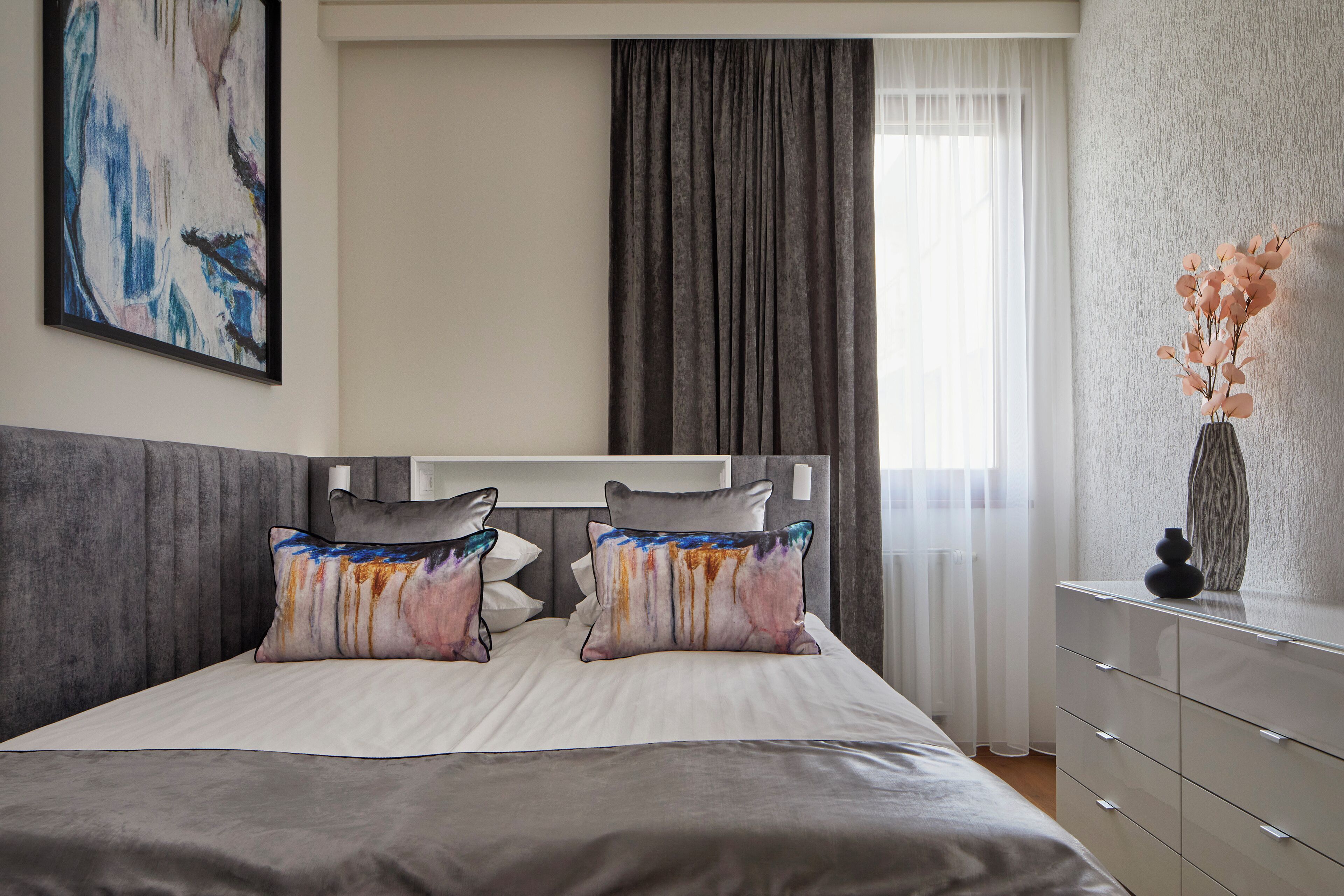 Une chambre à coucher moderne avec un grand lit, deux coussins décoratifs, une table de chevet et un vase rempli de fleurs.