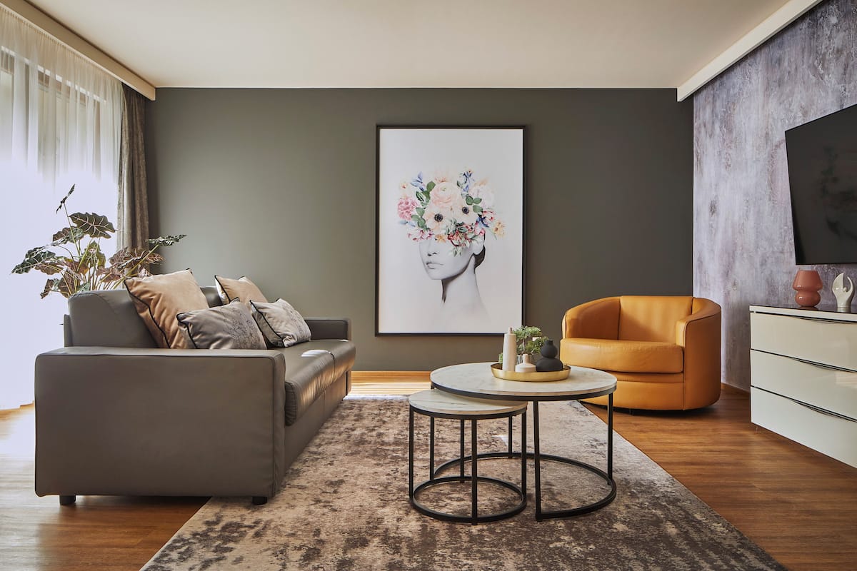 Ein modernes Wohnzimmer mit einem grauen Sofa, einem orangefarbenen Sessel, einem runden Couchtisch und einem großen, gerahmten Kunstwerk an der Wand.