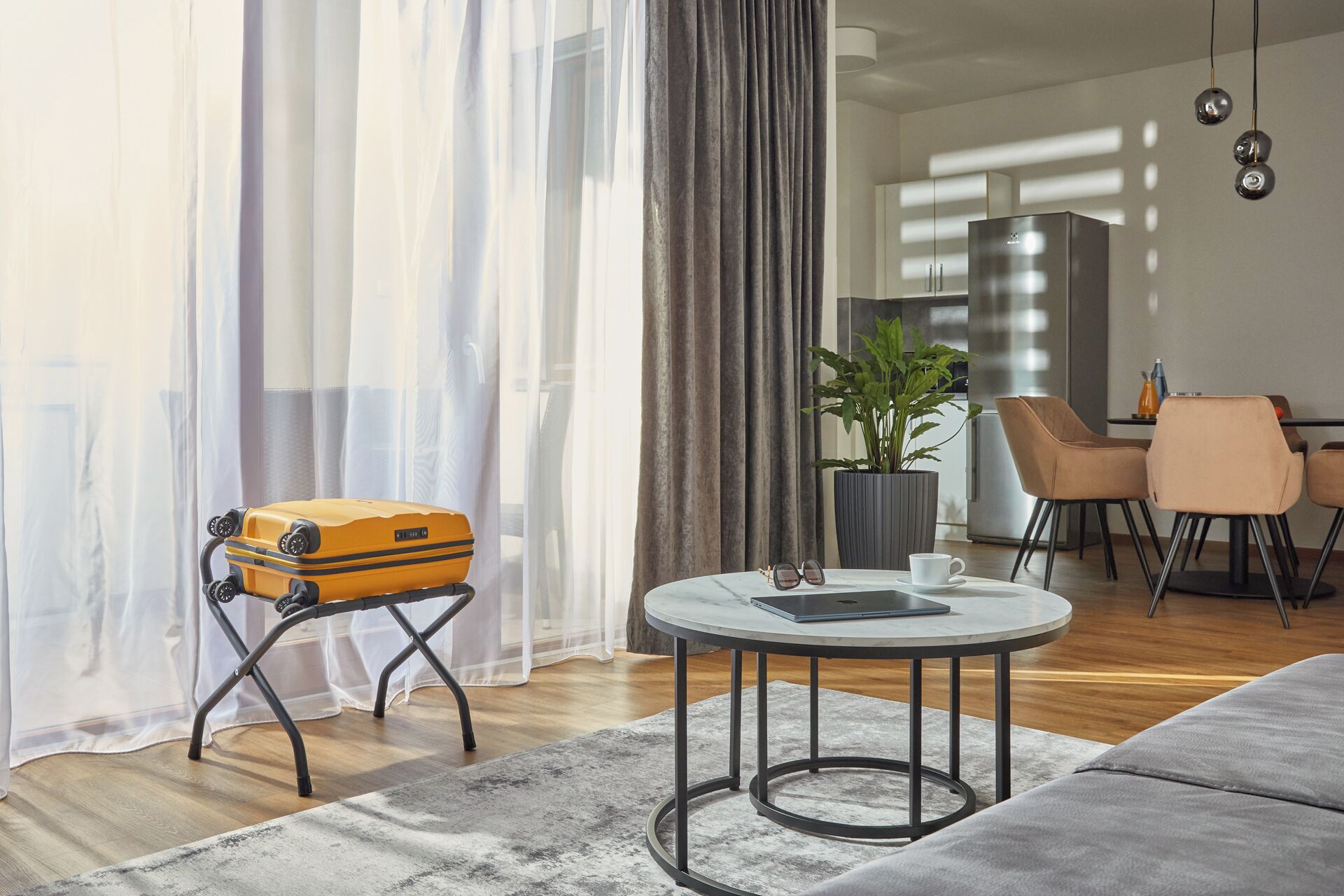 A modern living room with a sofa, a small round table with a cup and a tablet, a suitcase, and a plant.