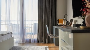 A modern hotel room with a bed, a desk with a laptop, a chair, a coffee maker, and a large window with curtains.