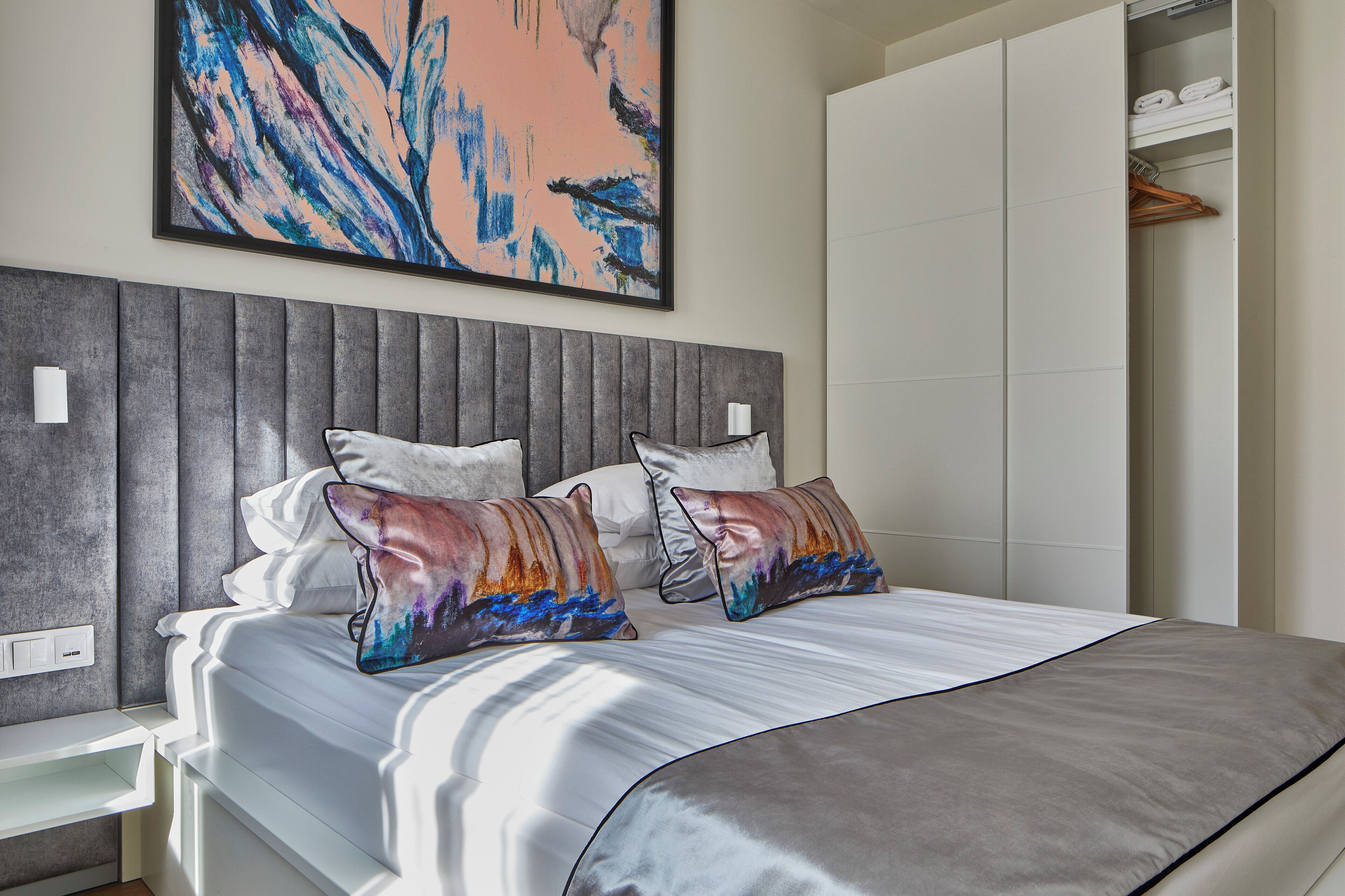 A modern bedroom with a large bed, a grey headboard, and a colorful abstract painting above it.