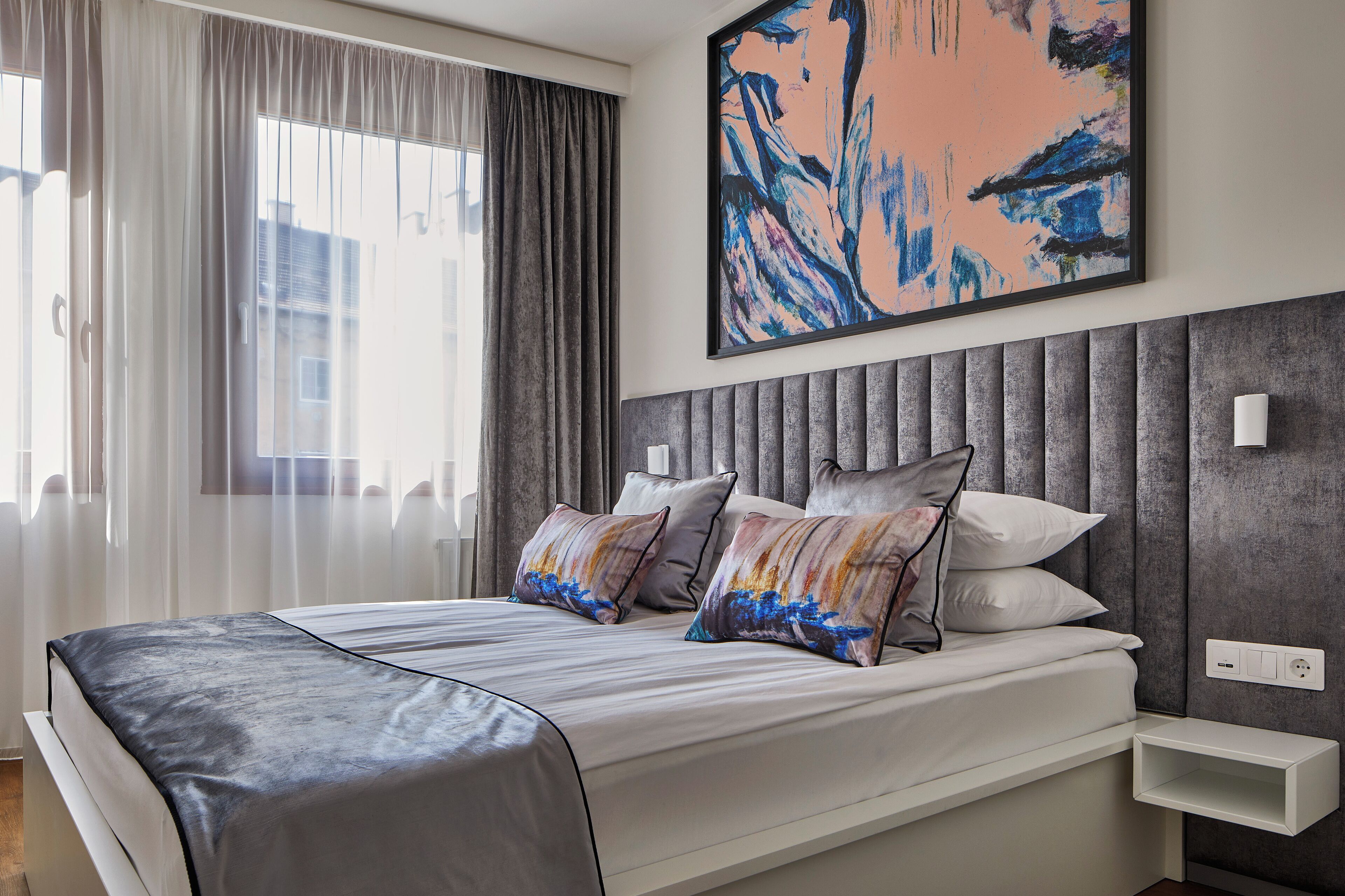 Quarto de hotel moderno com uma cama grande, cabeceira estofada cinza e um quadro em cima.