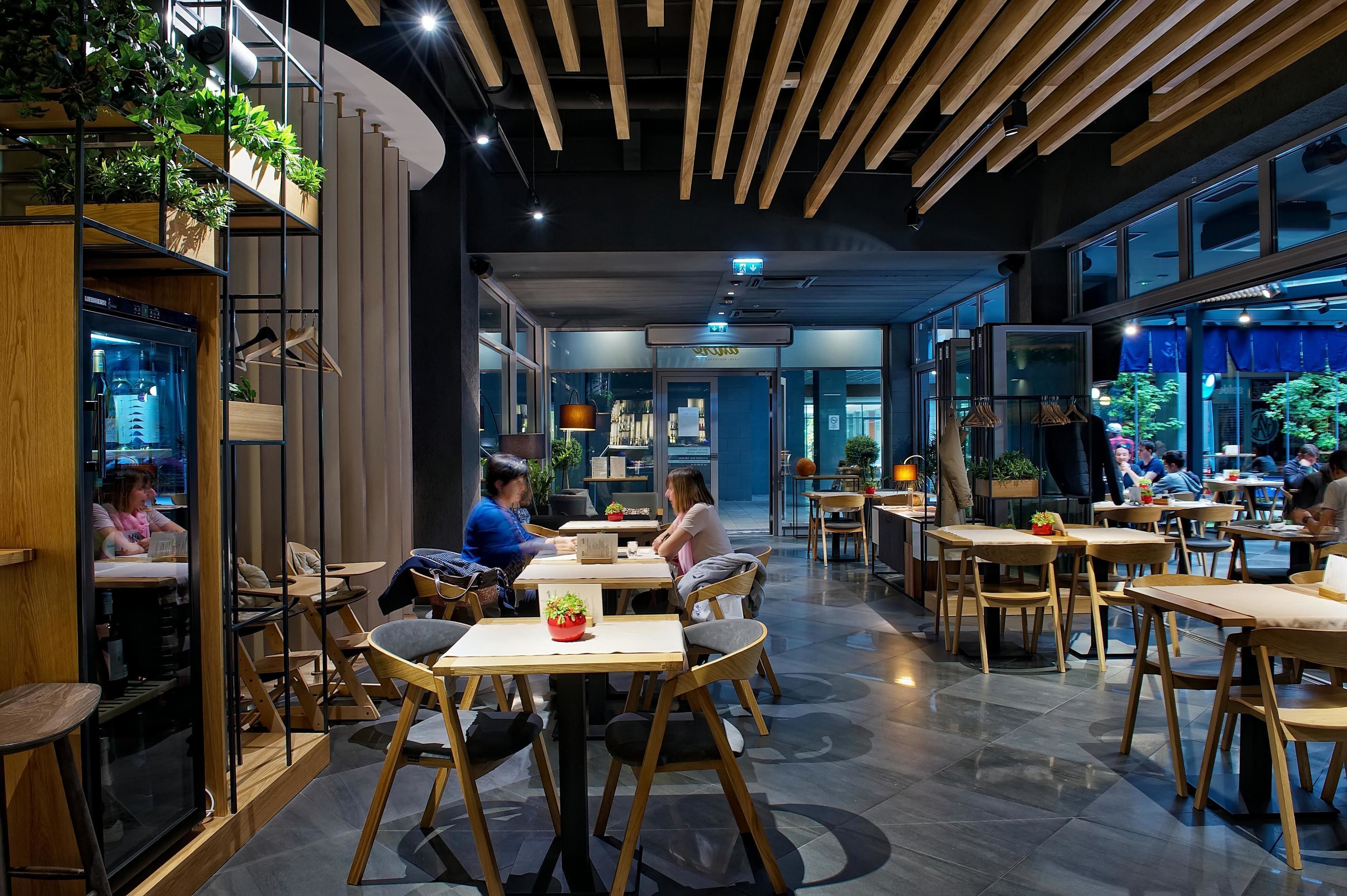 A modern restaurant with wooden furniture, large windows, and indoor plants.
