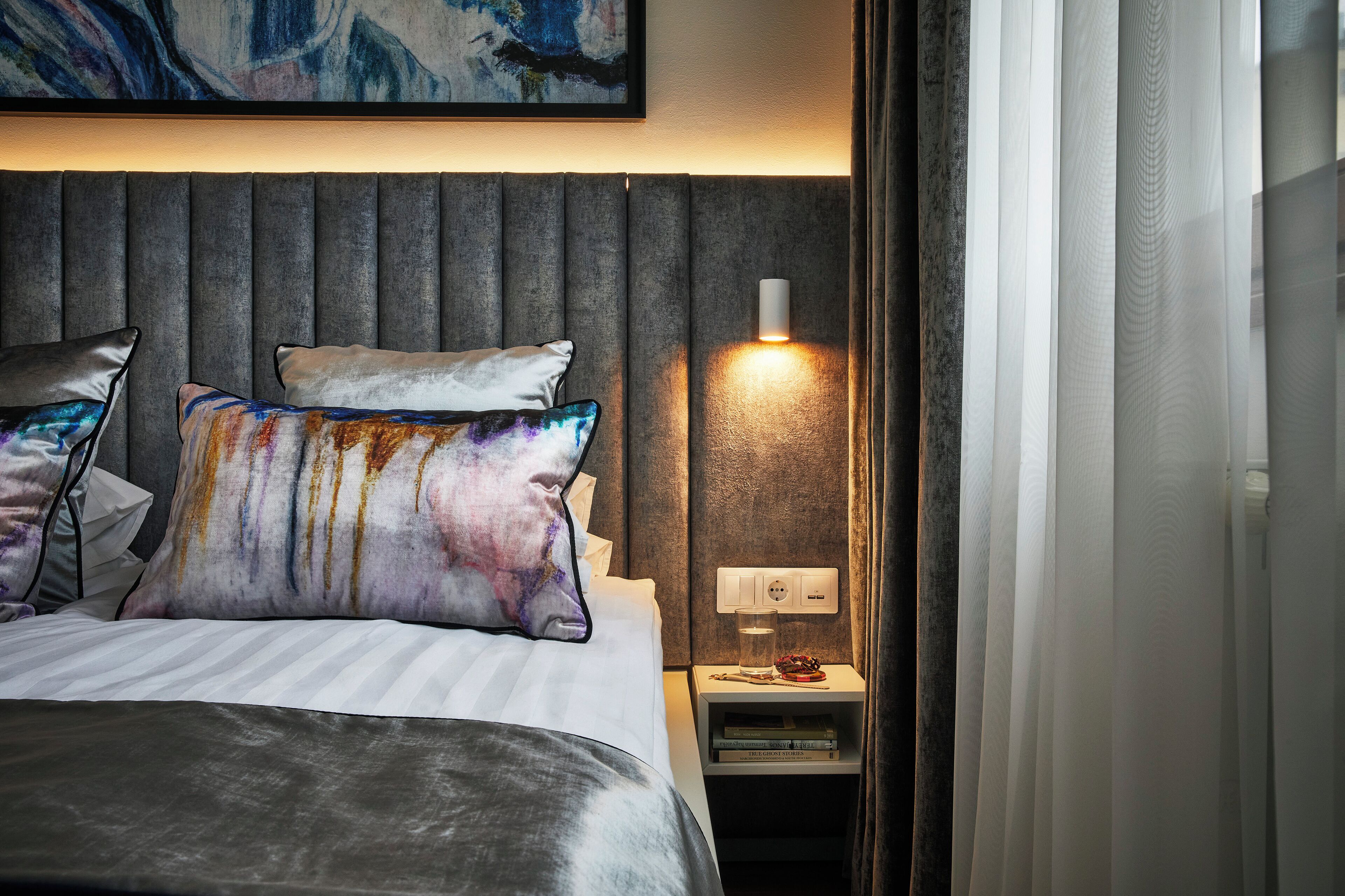 Un lit bien fait, avec une tête de lit grise, une table de chevet avec une lampe et un tableau au-dessus du lit.