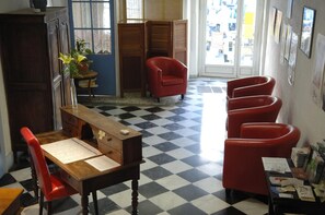 A room with a checkered floor, a desk with papers, a red chair, a red armchair, and a window with a view of the outside.