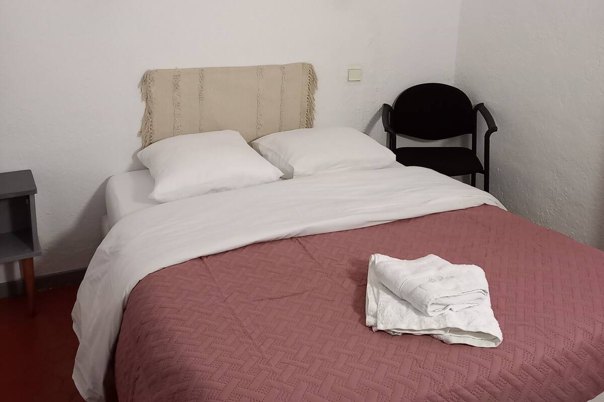 Un lit bien fait, avec du linge de lit blanc et une couverture rose, un miroir rond accroché au mur, une chaise noire et une petite table de chevet avec une lampe.