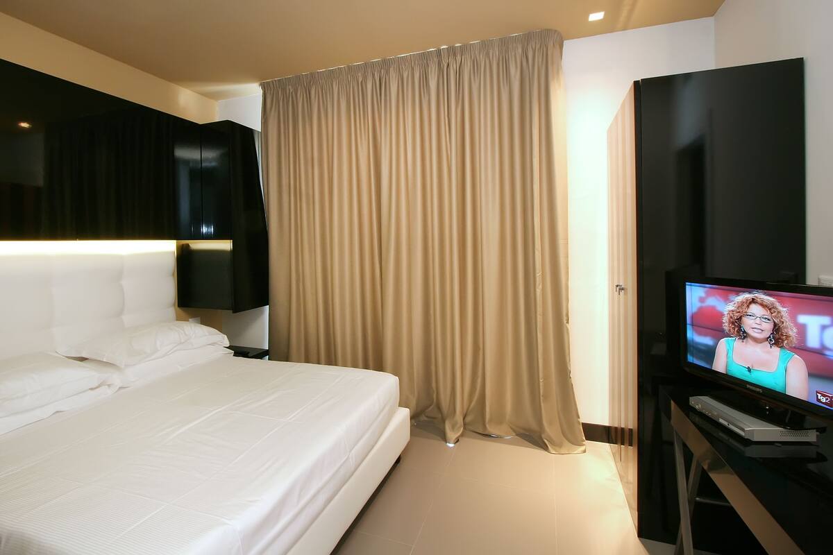 Quarto de hotel com uma cama grande, uma televisão em um suporte e uma cortina.