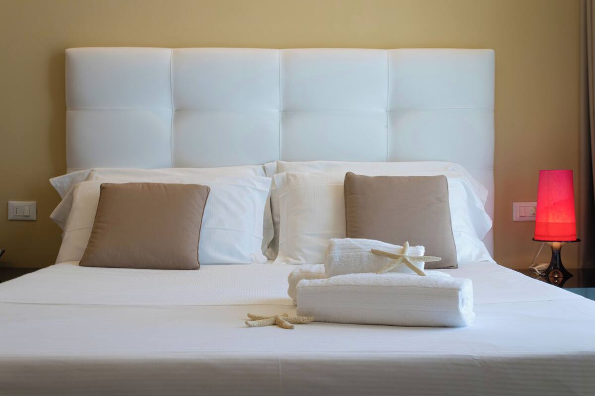 Cama bem arrumada com lençóis brancos e duas almofadas, um abajur vermelho sobre uma mesa de cabeceira, e uma toalha com um laço.