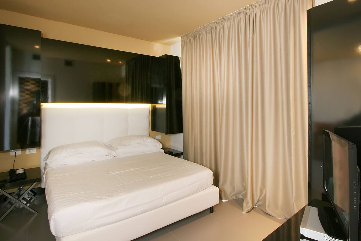 Quarto de hotel moderno com uma cama grande, uma televisão de tela plana e uma mesinha de cabeceira.
