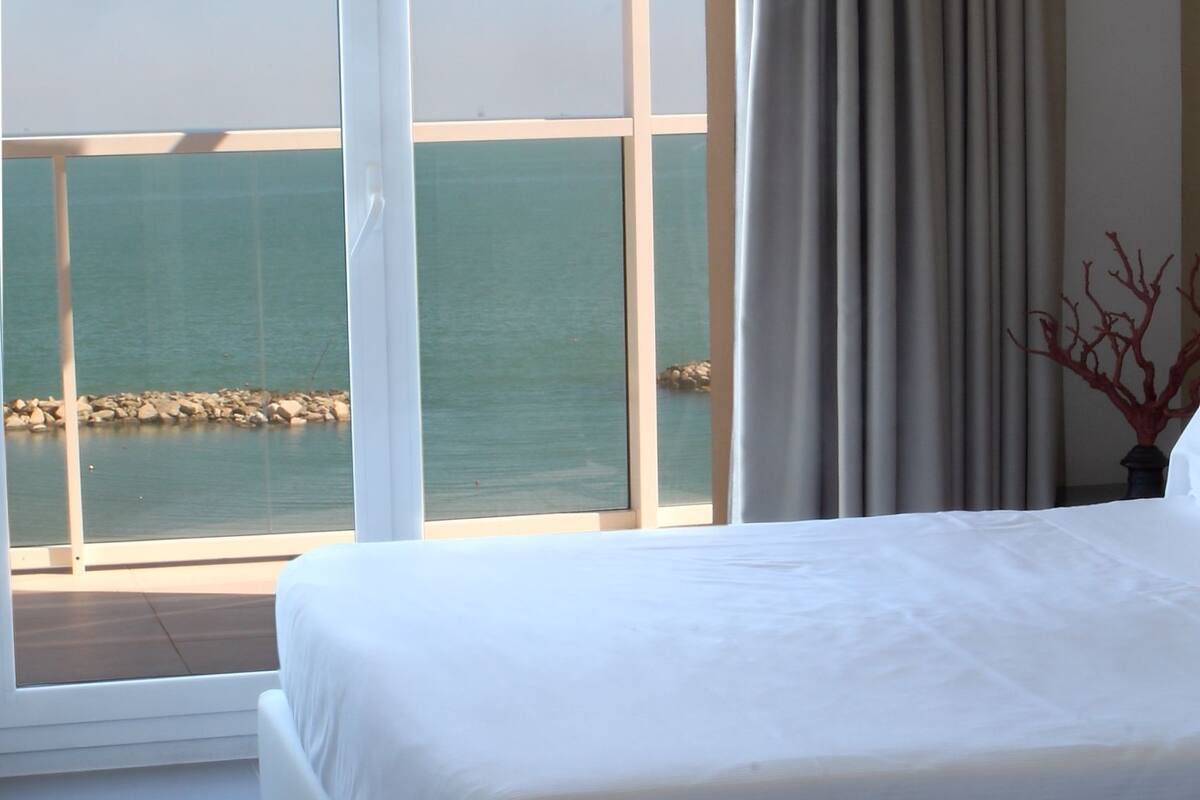 Quarto de hotel com uma cama grande, vista para o mar através de uma porta de correr de vidro e uma cortina na janela.