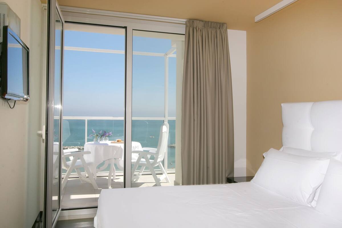 Quarto de hotel com cama, televisão, porta de vidro deslizante para varanda com mesa e cadeiras, e vista para o mar.