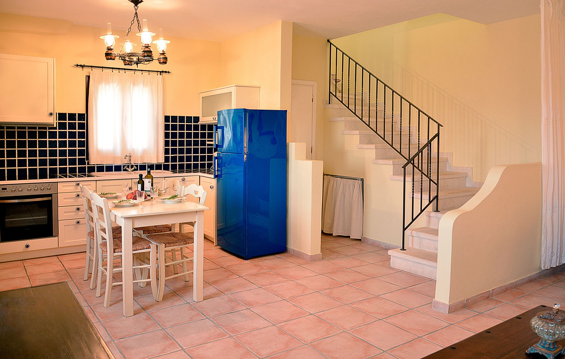 A kitchen with a dining table, chairs, a blue refrigerator, a chandelier, and a staircase.