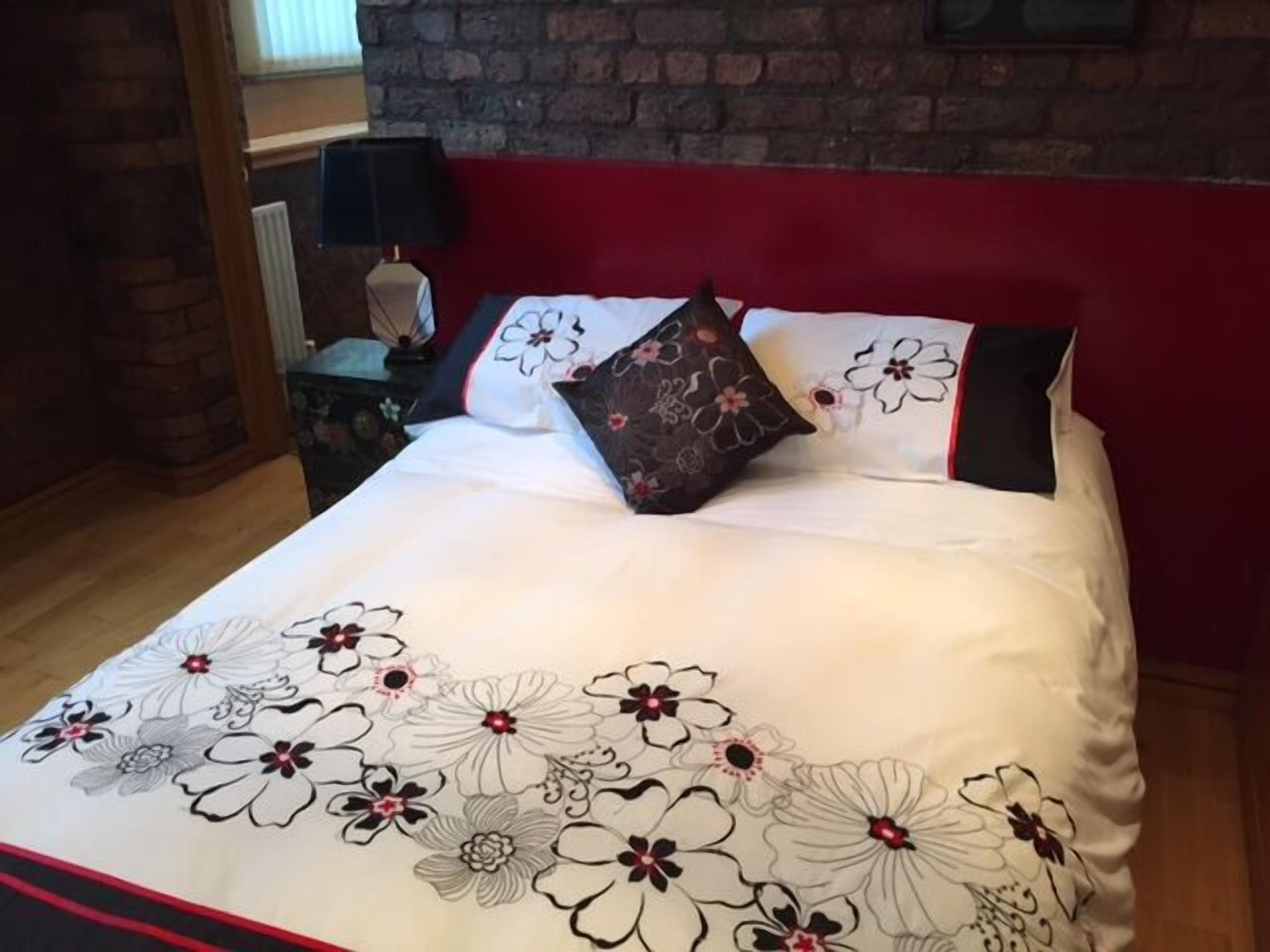Un lit bien fait, recouvert d’une couvre-lit à motifs floraux, avec une tête de lit rouge et une table de chevet sur laquelle se trouvent une lampe et un vase.