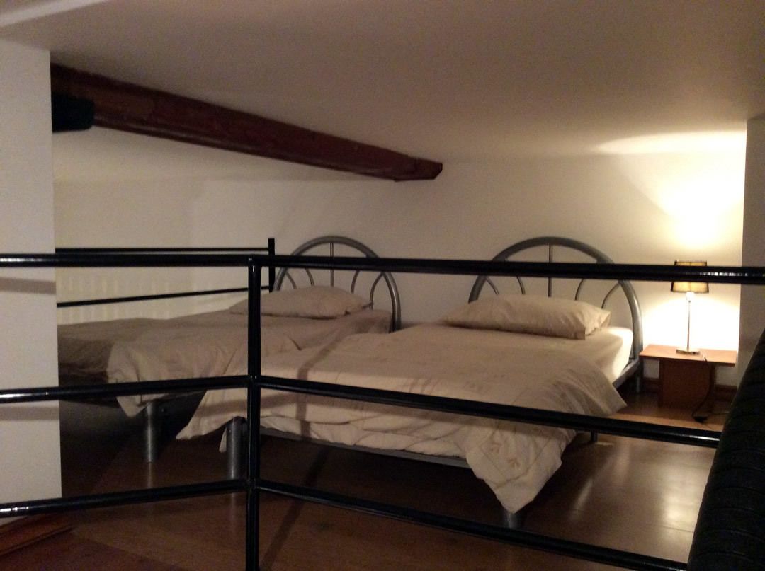A room with two beds, a wooden beam ceiling, a nightstand with a lamp, and a view of the outside.