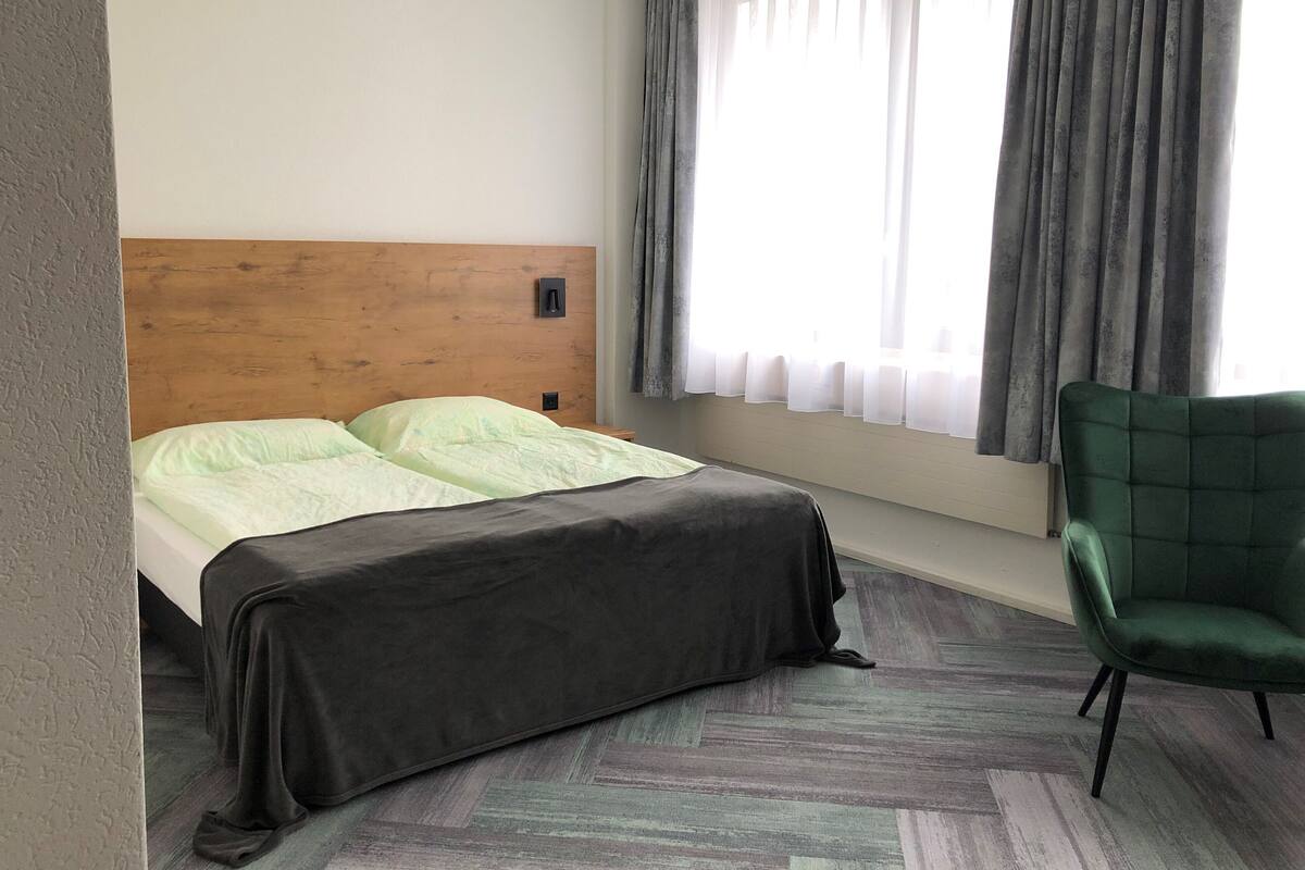 A hotel room with a wooden headboard, a bed with a grey bedspread, a green armchair, and a window with grey curtains.