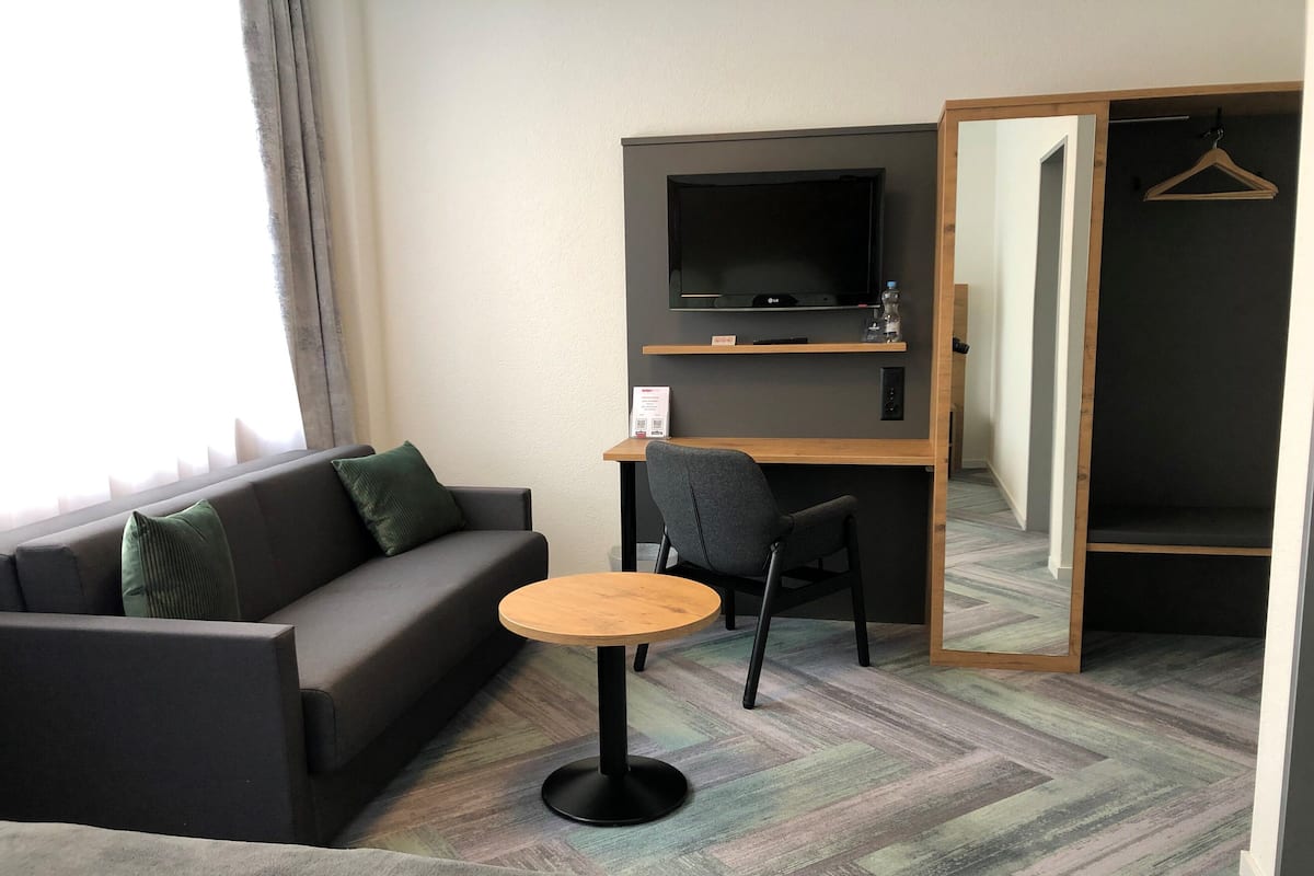 A hotel room with a sofa, a small round table, a TV mounted on the wall, and a wooden desk.
