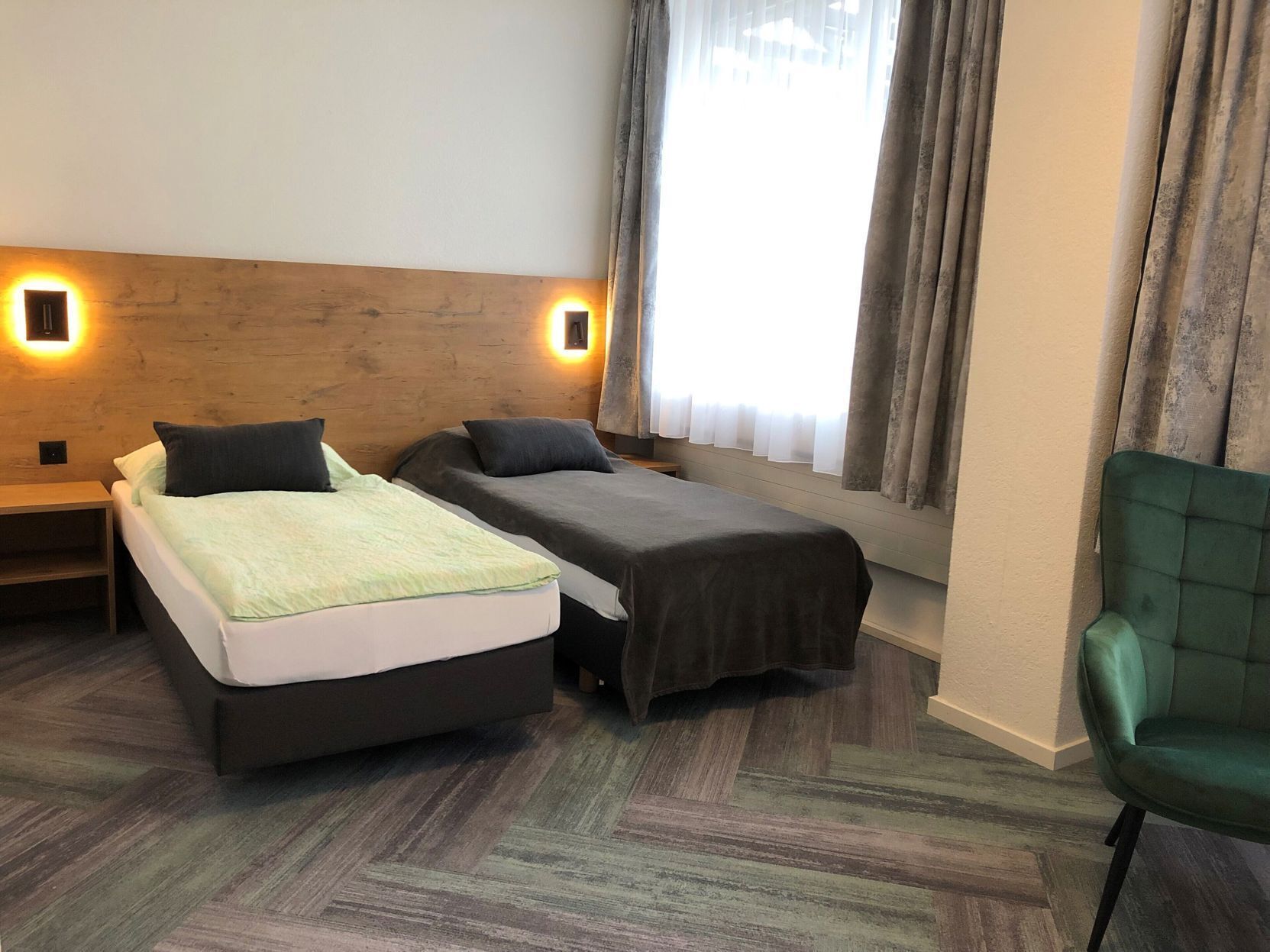 A hotel room with two beds, a green chair, and a window with curtains.