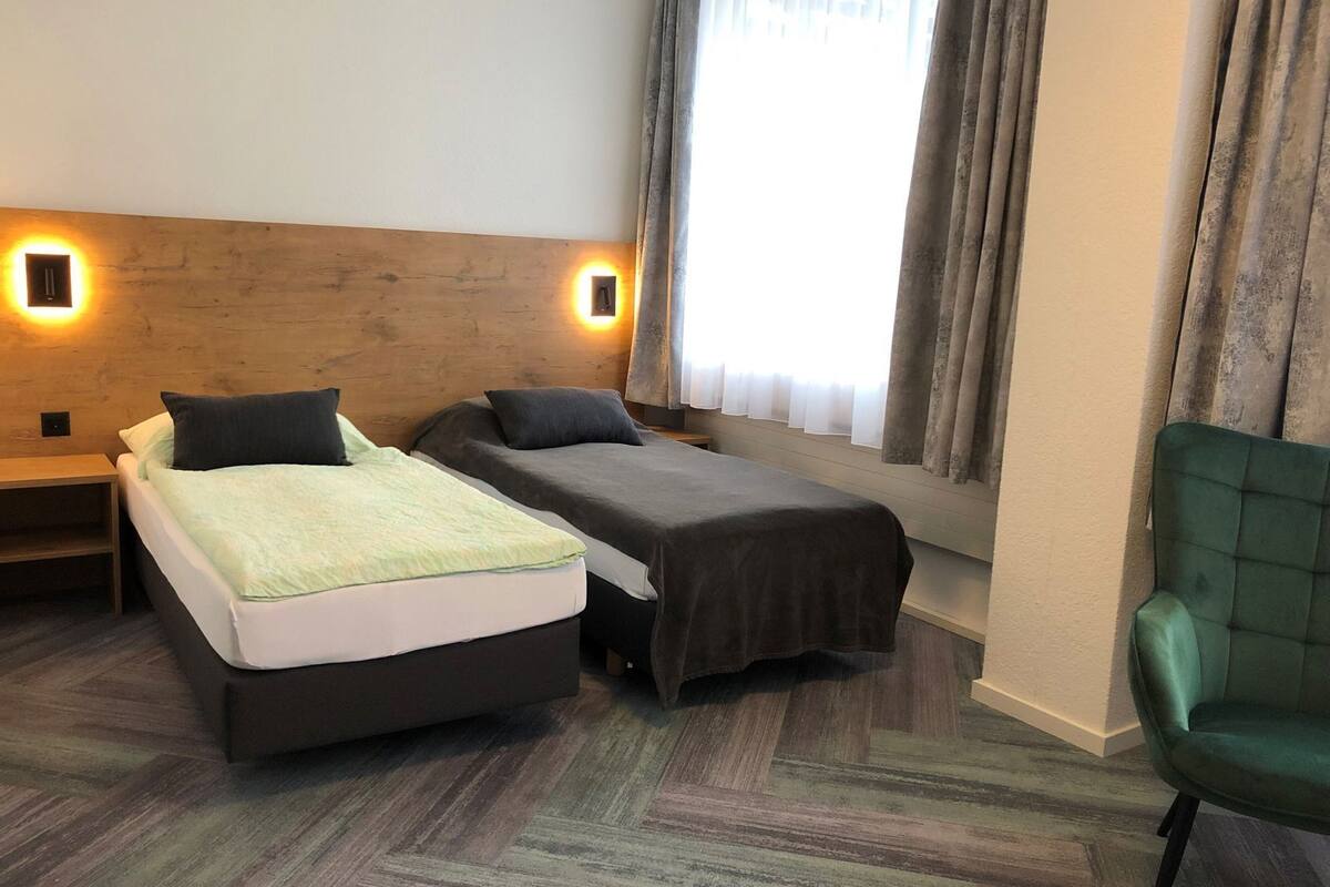 A hotel room with two beds, a green chair, and a window with curtains.